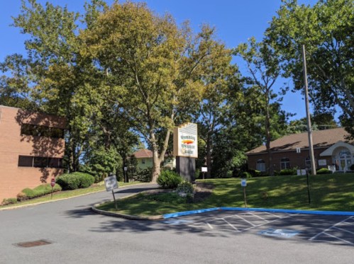 A sunny day at a facility entrance with trees, a sign, and a parking area.
