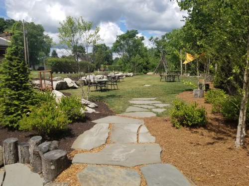 A stone pathway leads through a green park with trees, tables, and rocks under a partly cloudy sky.