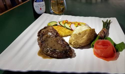 A plate featuring a steak with sauce, baked potato, omelet, vegetables, and smoked salmon, with a drink in the background.