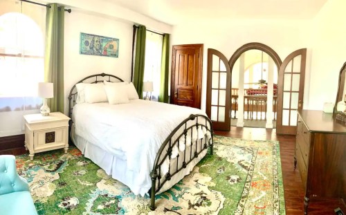 A bright bedroom featuring a metal bed, colorful rug, and large windows with green curtains.