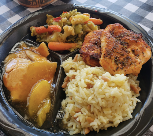 A black tray with rice, grilled chicken, sautéed vegetables, and sliced peaches in syrup.