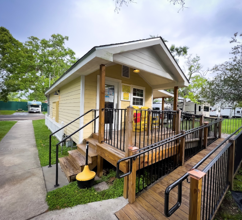 A small yellow house with a wooden porch and ramp, surrounded by greenery and a cloudy sky.
