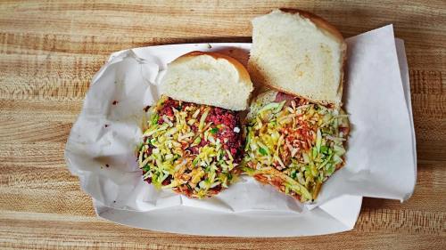 Two sandwiches on a paper tray, each topped with colorful shredded vegetables and served with a bun on the side.