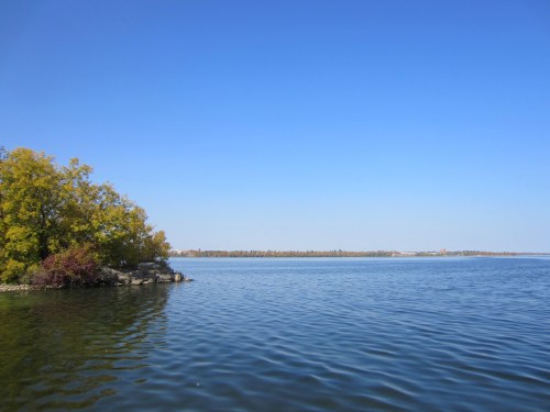 A serene lake scene with calm waters, a clear blue sky, and trees with autumn foliage along the shore.