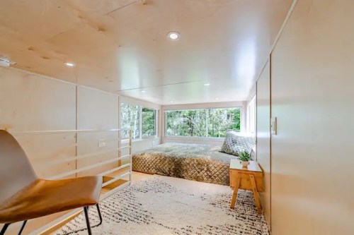 Cozy, modern bedroom with large windows, a bed, and minimalist decor in a serene, nature-filled setting.
