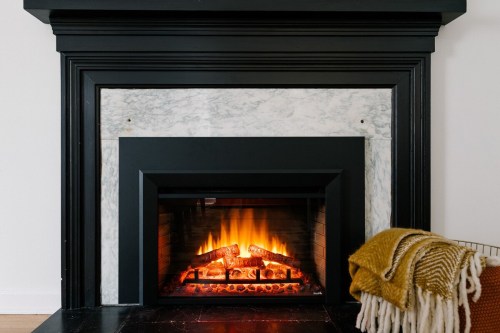 A cozy fireplace with glowing flames, framed by a black and marble mantel, beside a warm, knitted throw blanket.