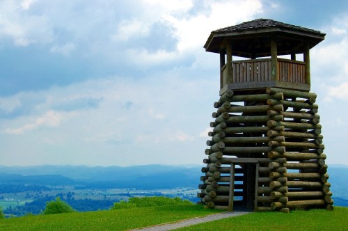 A wooden observation tower on a grassy hill, overlooking a valley with distant mountains under a cloudy sky.