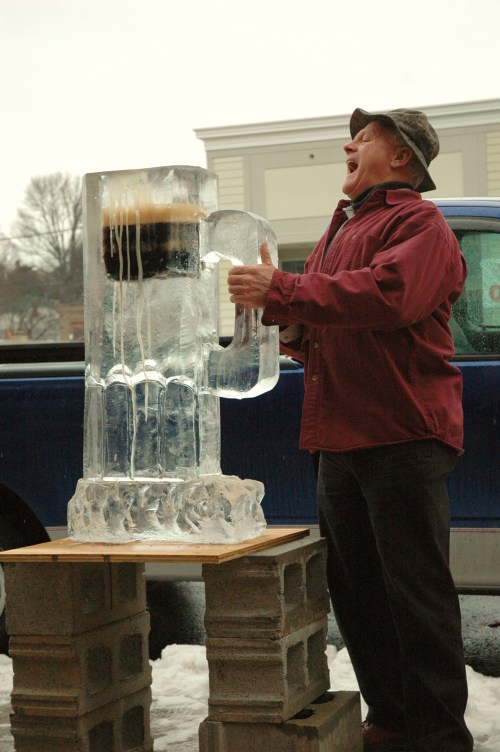 A man in a red jacket joyfully poses with a large ice sculpture of a beer mug outdoors.