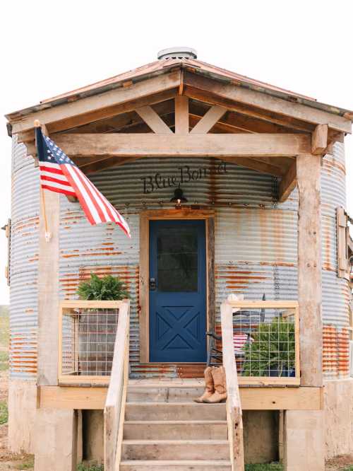 A rustic round building with a blue door, wooden porch, and an American flag, surrounded by greenery.