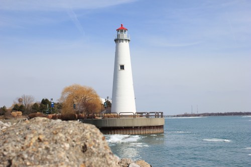 A tall white lighthouse with a red top stands by the water, surrounded by rocky shore and trees under a clear sky.