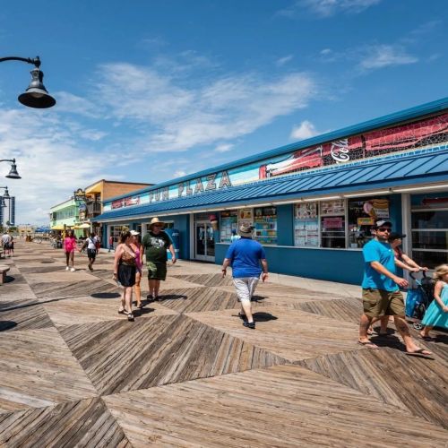 A busy boardwalk scene with people walking past colorful shops under a clear blue sky.