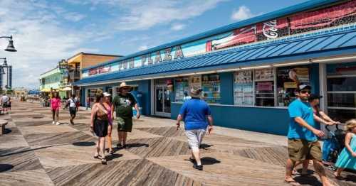 People walking along a boardwalk near a colorful shop called "Fun Plaza" on a sunny day.