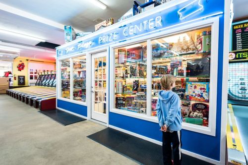 A child stands in front of a brightly lit prize center filled with toys and games behind glass windows.