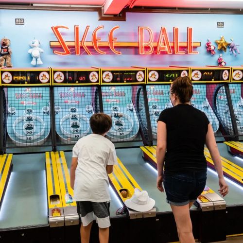 Two children play skee ball at an arcade, with colorful lights and stuffed animals displayed above the lanes.