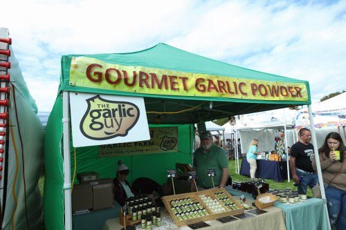 A green tent labeled "Gourmet Garlic Powder" with products displayed and people browsing at a market.