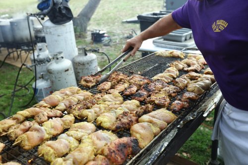 A person grilling marinated chicken on a barbecue, with smoke and outdoor cooking equipment in the background.