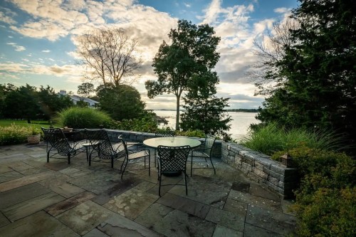 A serene patio with a table and chairs overlooking a lake, surrounded by trees and a cloudy sky.