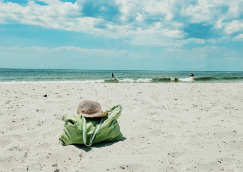 A green beach bag and a straw hat sit on sandy shore, with gentle waves and a cloudy sky in the background.
