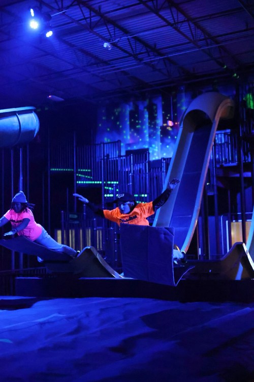 Two people in neon shirts jump off a trampoline into a dark, colorful indoor play area with slides.