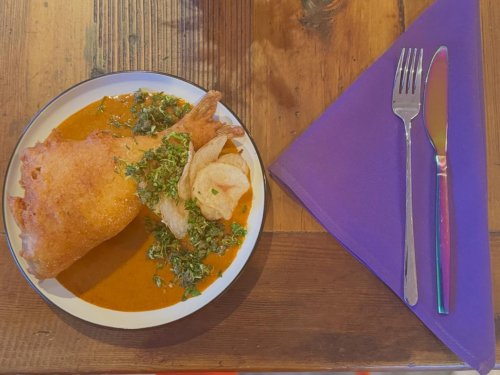 A plate with fried fish, a rich sauce, and garnished greens, accompanied by a purple napkin and silverware.