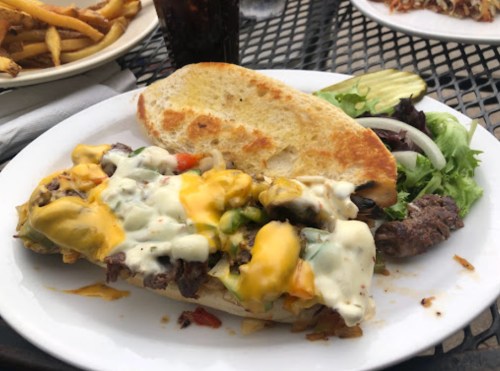 A loaded sandwich with melted cheese, grilled meat, and vegetables, served with a side of salad and pickles.