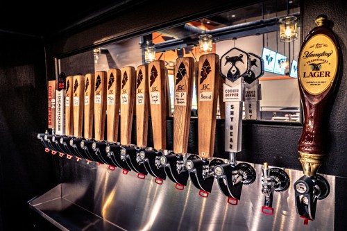 A row of wooden beer taps with various labels, set against a sleek metal backdrop in a bar.
