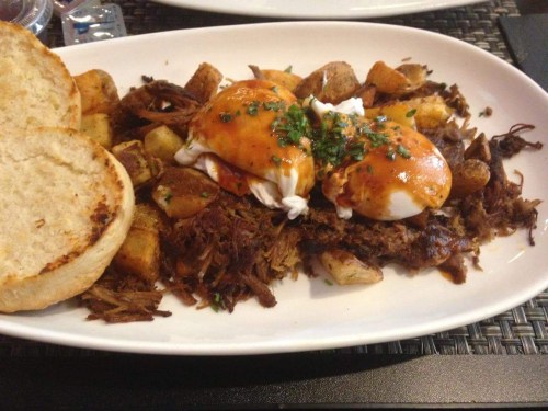 A plate of pulled beef with poached eggs, seasoned potatoes, and a toasted bun, garnished with herbs.