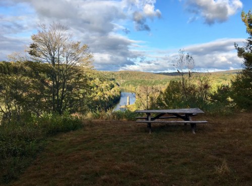 A scenic view of a river surrounded by trees, with a picnic table in the foreground under a partly cloudy sky.