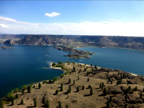 Aerial view of a serene lake surrounded by hills and greenery under a clear blue sky.