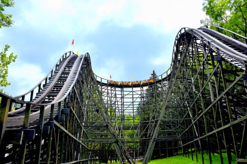 A wooden roller coaster with steep hills and a cloudy sky in the background, surrounded by trees.