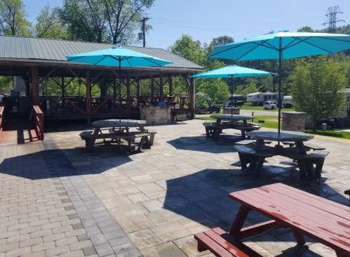 Outdoor seating area with picnic tables and large blue umbrellas, surrounded by greenery and a pavilion in the background.