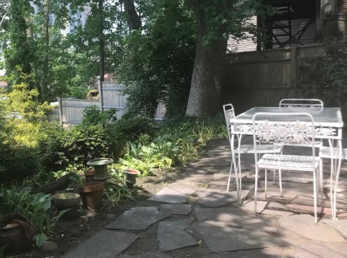 A serene garden scene featuring a stone path, potted plants, and a white metal table with chairs under leafy trees.