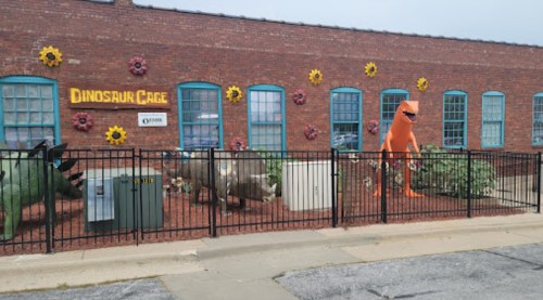Colorful dinosaur sculptures and a large orange dinosaur figure outside a brick building with flower decorations.