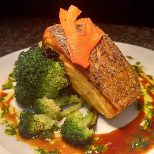 A beautifully plated salmon fillet topped with a carrot slice, served with steamed broccoli and a drizzle of sauce.