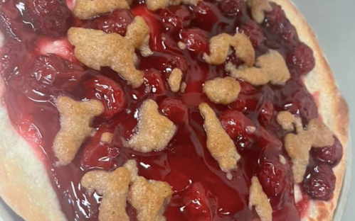 A cherry pie topped with a glossy red cherry filling and scattered cookie-like pieces on top.