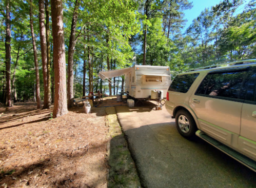 A camper parked under trees with an awning extended, next to a silver SUV on a gravel path.