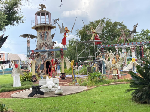 A whimsical outdoor sculpture garden featuring a lighthouse, angels, and various artistic figures amidst greenery.