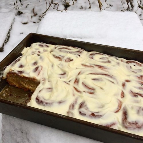 A tray of cinnamon rolls with cream cheese frosting sits on a snowy surface. One roll is partially cut.
