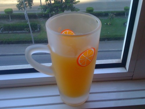A frosted glass mug filled with a yellow beverage, sitting on a windowsill with a view of greenery outside.