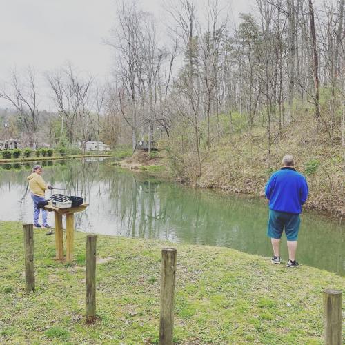 Two men fishing by a calm pond surrounded by trees, one standing on the bank and the other at a nearby table.