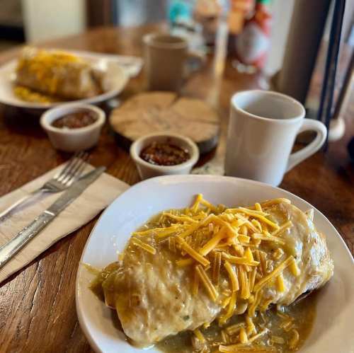 Two large burritos topped with cheese, served with small bowls of salsa and a coffee cup on a wooden table.