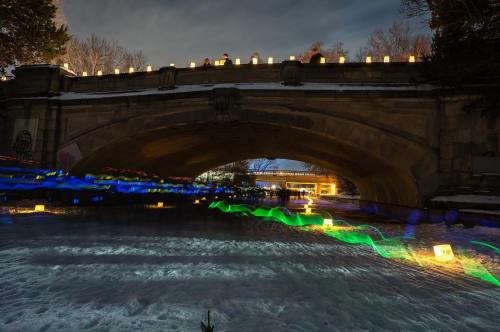 A bridge illuminated at night, with colorful lights reflecting on a snowy river below. Lanterns line the bridge.