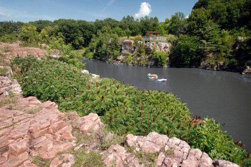 A serene river scene with rocky banks, lush greenery, and small boats floating on the water under a clear blue sky.