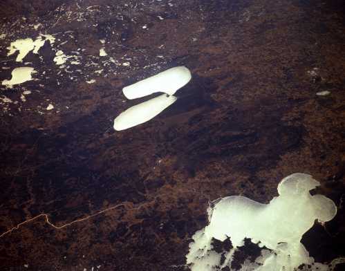 Aerial view of a landscape with large ice formations on dark terrain, resembling abstract shapes against the earth.