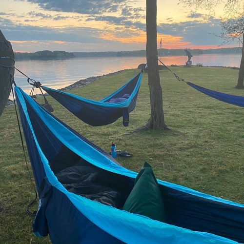 Two blue hammocks are set up by a lake at sunset, with a grassy area and trees in the background.