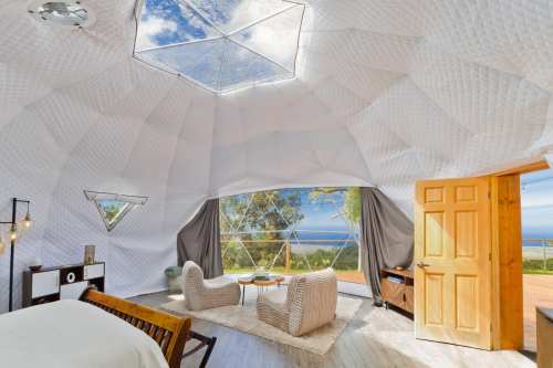 A bright, modern geodesic dome interior with panoramic windows, featuring cozy seating and scenic ocean views.