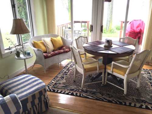 Cozy sunroom with a round table, four chairs, a wicker couch, and a lamp, surrounded by large windows and greenery.