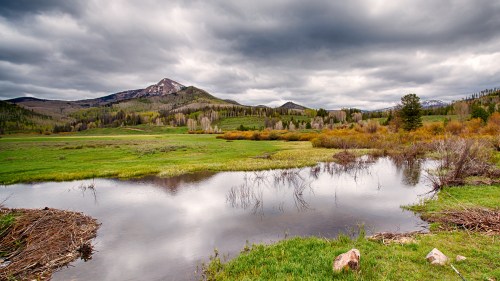 A serene landscape featuring a calm pond, lush green fields, and mountains under a cloudy sky.
