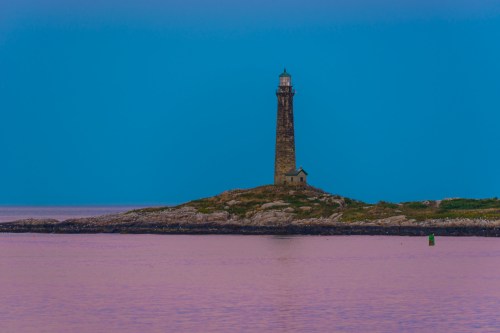 A tall lighthouse stands on a rocky island, surrounded by calm waters under a blue sky at dusk.