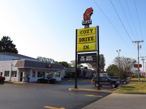 A drive-in restaurant with a large sign reading "Cozy Drive In" and "We Have Kids Meals," surrounded by parked cars.